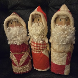 May include: Three handcrafted fabric Santa Claus figurines featuring quilted red and white patterns. Each figure wears a hooded cloak with a cream-colored trim, a curly white wool beard, and a hand-painted face with simple black eyes.