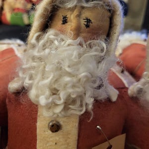 May include: A handmade Santa Claus doll with a red felt coat, cream-colored trim, and a fluffy white beard. The doll has stitched eyes and rosy cheeks. A small tag is attached.