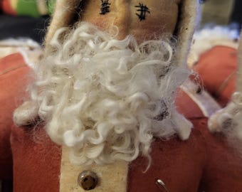 May include: A handmade Santa Claus doll with a red felt coat, cream-colored trim, and a fluffy white beard. The doll has stitched eyes and rosy cheeks. A small tag is attached.