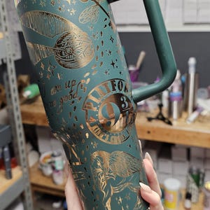 May include: Teal travel mug with gold accents and a handle. The mug features gold illustrations of a feather, stars, and a winged creature. Text on the mug includes "Platform 9 3/4" and "I am up to no good."