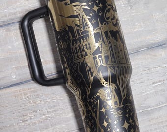 May include: Black travel mug with a gold-colored design featuring a castle, stars, and wands. The mug has a black handle and a black lid with a gold band. The design is etched into the mug's surface.