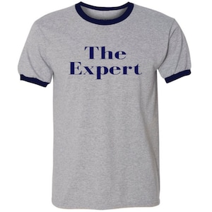 May include: A gray t-shirt with a navy blue ringer. The shirt has the text "The Expert" printed on the front in navy blue.