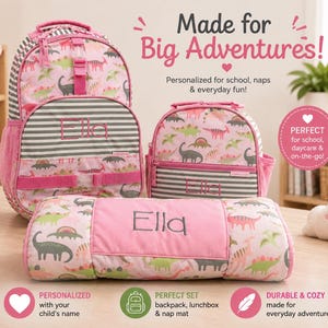 Op de afbeelding: Roze rugzak, lunchbox en slaapmat met dinosauruspatroon. Gepersonaliseerd met de naam "Ella". Met de tekst "Made for Big Adventures!" en "Perfect for school, daycare & on-the-go!"