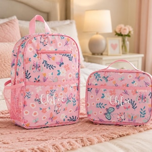 Könnte beinhalten: Ein rosa Rucksack und eine passende rechteckige Lunchtasche mit einem bunten Blumen- und Schmetterlingsmuster. Jeder Artikel ist mit dem Namen Chloe in weißer Schreibschrift personalisiert und verfügt über eine Reißverschlusstasche vorne.