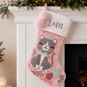 Puede incluir: Un calcetín navideño rosa con un borde blanco que lleva el nombre Lola bordado. El calcetín muestra un gato gris con corona, un ovillo de lana rojo y un regalo plateado. Dos pompones rosas están sujetos al lazo superior para colgarlo.