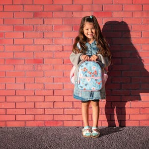 May include: A young girl stands in front of a red brick wall, holding a light blue backpack with a floral design. The backpack has a pink zipper and straps. She is wearing a denim dress, a gray sweater, and sandals.
