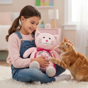 May include: A young girl holds a pink cat plush toy with a pink ribbon and the name "Ruby" embroidered on it. An orange tabby cat reaches toward the plush toy. The girl is wearing a pink sweater and denim overalls.