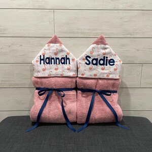 Two pink towels with names "Hannah" and "Saddie" printed on them, each with a blue ribbon tied around the center.