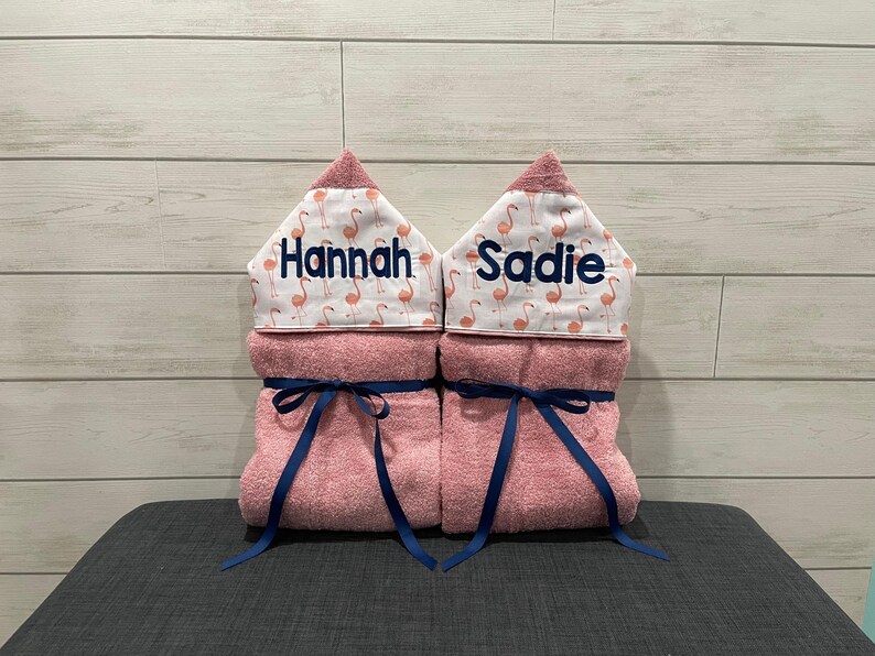 Two pink towels with names "Hannah" and "Saddie" printed on them, each with a blue ribbon tied around the center.