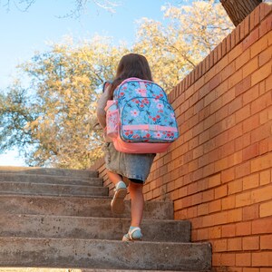 May include: A child ascends concrete steps, carrying a light blue backpack adorned with a floral pattern and pink accents. The backpack has a matching water bottle holder. A brick wall and tree branches frame the scene under a clear blue sky.