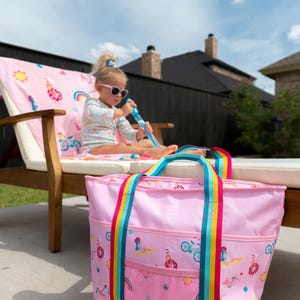 A young girl sits on a pink chair with a pink tote bag next to her, in an outdoor setting with a wooden fence and a house in the background.