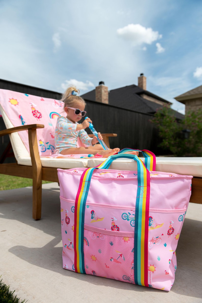 A young girl sits on a pink chair with a pink tote bag next to her, in an outdoor setting with a wooden fence and a house in the background.
