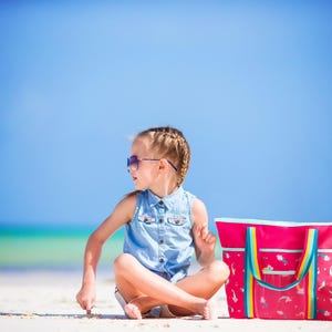 A young girl in a denim dress sits on a sandy beach, wearing sunglasses and accompanied by a pink beach bag and an orange towel.