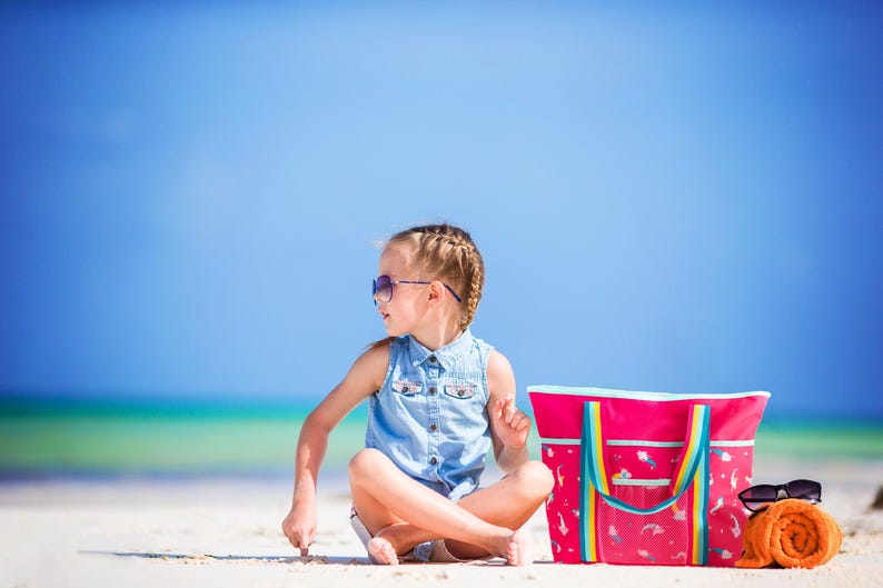 A young girl in a denim dress sits on a sandy beach, wearing sunglasses and accompanied by a pink beach bag and an orange towel.