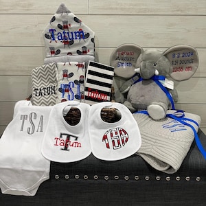 Puede incluir: Una canasta de regalo para bebés con una variedad de artículos. Incluye un peluche de elefante gris, una toalla con capucha con un patrón de castor, baberos, una manta y un body. Todos los artículos están personalizados con el nombre "Tatum" e iniciales.