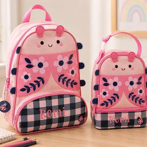 May include: Pink backpack and matching lunch bag set. Both feature a ladybug design with floral accents and a gingham bottom. The backpack and lunch bag have the name "Remi" printed on them.