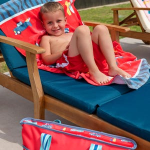 A young boy is sitting on a blue lawn chair, smiling and wearing red swim trunks. There is a red and blue bag next to the chair.