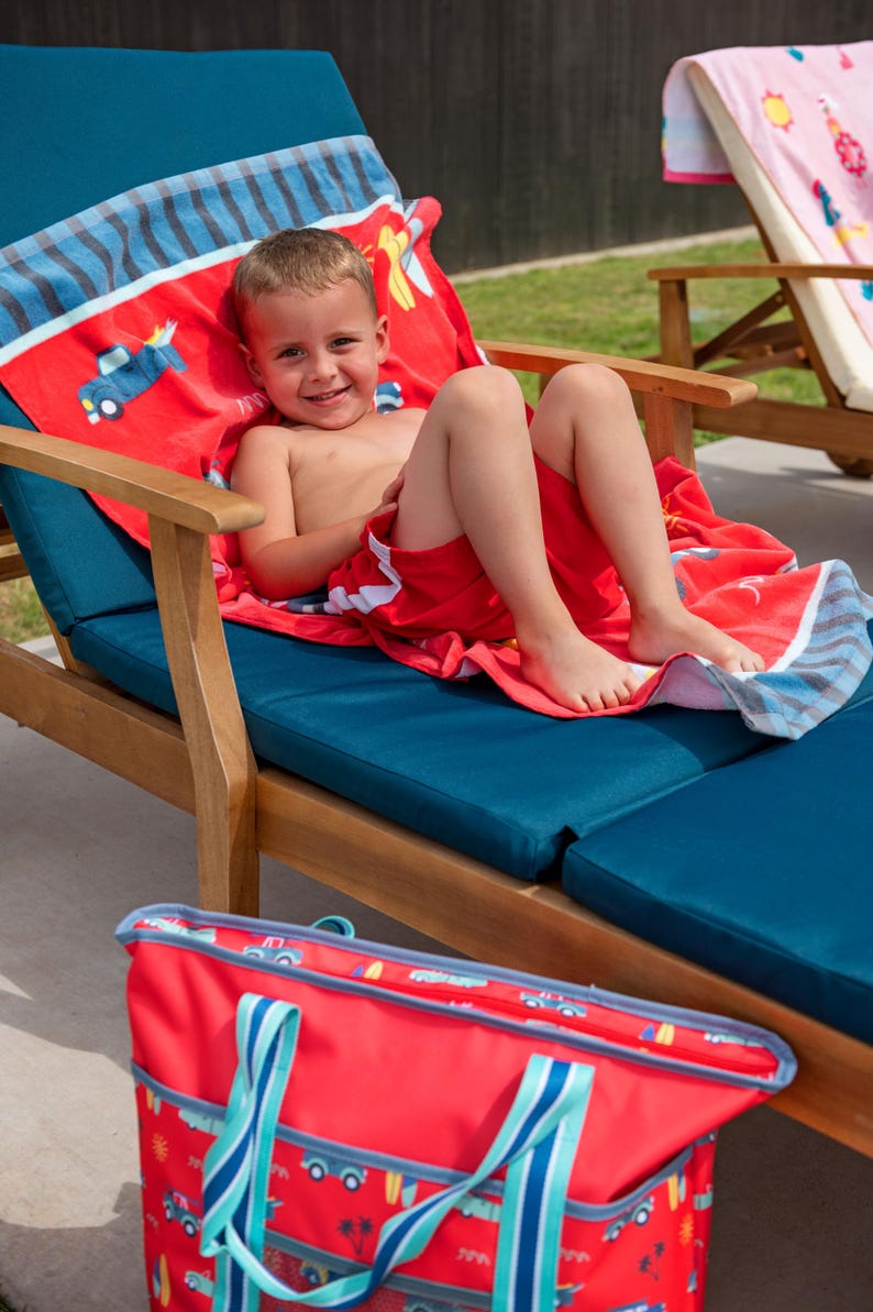 A young boy is sitting on a blue lawn chair, smiling and wearing red swim trunks. There is a red and blue bag next to the chair.