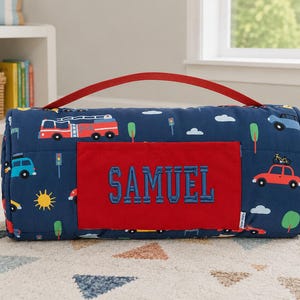May include: A rolled-up navy blue blanket with a red handle. The blanket features a pattern of cars, fire trucks, trees, and clouds. A red patch with the name "SAMUEL" in blue letters is sewn on the front.