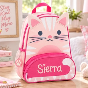 May include: A light pink backpack featuring a cat face design with pointed ears, dark whiskers, and a pink nose. The front pocket is a darker shade of pink with the name Sierra printed in white, accompanied by a small paw print zipper pull charm.