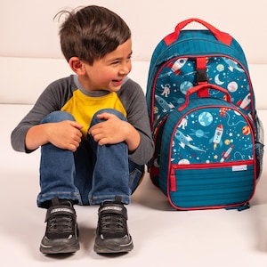 May include: A teal and red backpack and matching lunch bag set. The bags feature a space-themed design with rockets, planets, and astronauts. The backpack has a top handle and adjustable straps. The lunch bag has a front zipper pocket.