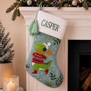 Può includere: Una calza di Natale con un design a dinosauro. La calza è azzurra con un dinosauro verde che indossa un maglione rosso e pattini. La parte superiore è bianca con il nome "CASPER" ricamato in verde.