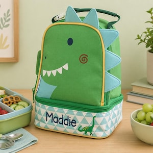 Könnte beinhalten: Grüne Lunchtasche im Dinosaurier-Design mit blauen Akzenten und dem Namen "Maddie" auf der Vorderseite. Die Tasche hat einen oberen Griff und ein unteres Fach mit geometrischem Muster. Eine passende Lunchbox und Besteck sind ebenfalls zu sehen.