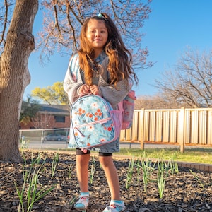 May include: A young person stands outdoors, wearing a denim jacket and carrying a light blue backpack with a floral design. The backpack has a pink pocket and a matching water bottle. The person is wearing shorts and sandals.