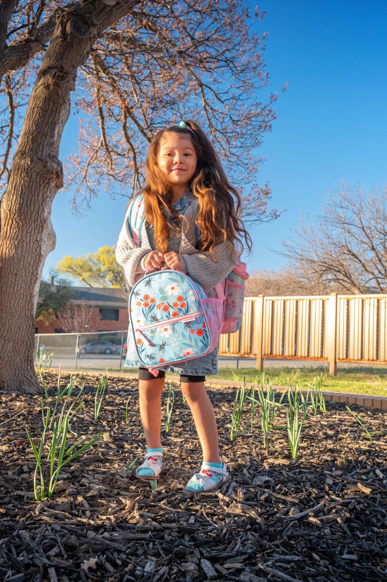 May include: A young person stands outdoors, wearing a denim jacket and carrying a light blue backpack with a floral design. The backpack has a pink pocket and a matching water bottle. The person is wearing shorts and sandals.