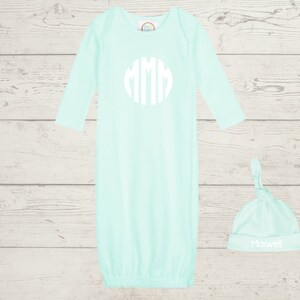 May include: A light blue baby gown with long sleeves and a white monogram on the front. A matching light blue knotted hat with the name "Maxwell" embroidered in white.