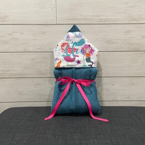 A towel with a mermaid design and a pink ribbon tied around it, placed on a gray surface against a wooden background.