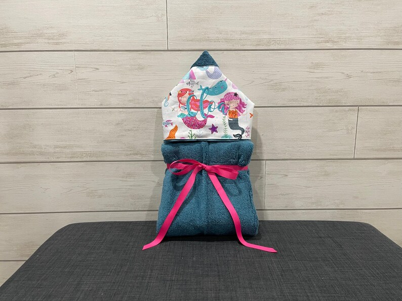 A towel with a mermaid design and a pink ribbon tied around it, placed on a gray surface against a wooden background.