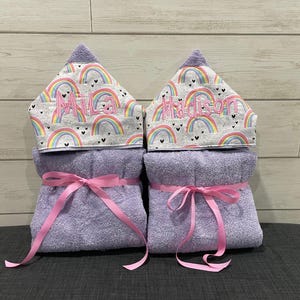 two purple towels with pink ribbons, each with a name written on it. The towels are placed on a gray surface against a wooden background.