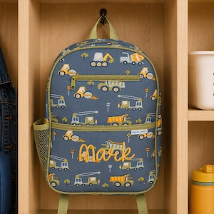 May include: A blue backpack featuring a repeating pattern of yellow and green construction vehicles, including dump trucks and cranes. The bag has a front zippered pocket, a side mesh pocket, and the name Mark embroidered in yellow cursive script.