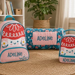 May include: A pink backpack, lunch bag, and pillow set with a floral design. The backpack and lunch bag have a red top with a white flower design and the name "Adeline" embroidered on them. The pillow is blue with a floral pattern and the name "Adeline".