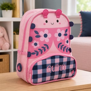 Op de afbeelding: Roze en blauwe kinderrugzak met een lieveheersbeestjesontwerp. De rugzak heeft een roze body met een lieveheersbeestjesgezicht, bloemenaccenten en een blauw-roze geruit vak met de naam "Quinn".