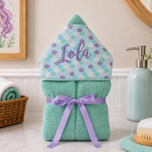 Pode incluir: Uma toalha com capuz turquesa com uma fita roxa e o nome "Lola" no capuz. O capuz apresenta um padr&atilde;o de escamas de sereia em tons de azul e roxo. Um patinho de borracha amarelo e uma imagem de sereia est&atilde;o no fundo.