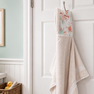 Pode incluir: Uma toalha de banho com capuz bege para beb&eacute; est&aacute; pendurada num gancho prateado numa porta branca. O capuz apresenta um padr&atilde;o floral com detalhes em rosa, p&ecirc;ssego e verde. Uma moldura com uma baleia e um cesto com um patinho de borracha s&atilde;o vis&iacute;veis.