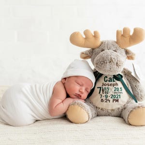 May include: A newborn baby wrapped in a white blanket and wearing a white cap, sleeping next to a plush moose toy. The moose has tan antlers and a green ribbon. Text on the moose reads: Cal Joseph, 7 lbs 5 oz, 20.5 inches, 7-9-2021, 8:24 PM.