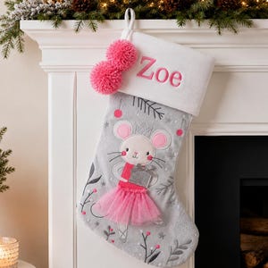 May include: A gray Christmas stocking with a white cuff embroidered with the name Zoe in pink. The stocking features a mouse illustration wearing a pink tutu and holding a silver gift, accented with two pink pom-poms and winter-themed embroidery.
