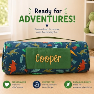May include: A rolled-up dark blue nap mat featuring a colourful dinosaur and tropical leaf pattern. A solid green rectangular patch on the front is embroidered with the name Cooper in yellow script. It includes a green carrying handle.