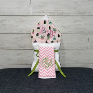 May include: A white hooded baby towel with a pink cactus print hood and a matching pink and white chevron washcloth. The washcloth has a green embroidered monogram. The towel is tied with a green ribbon.