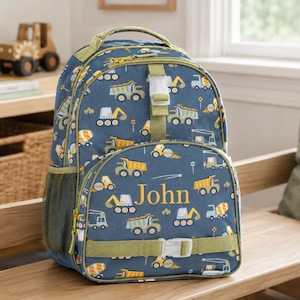 Könnte beinhalten: Ein blauer Rucksack mit einem Muster aus Baumaschinen, darunter Muldenkipper und Bagger. Der Name "John" ist gelb aufgedruckt. Der Rucksack hat grüne Akzente, Riemen und einen Griff. Eine Seitentasche ist sichtbar.