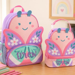 Könnte beinhalten: Ein rosa und lila Rucksack im Schmetterlings-Design und eine passende Lunchtasche. Der Rucksack hat ein großes Schmetterlingsflügel-Design mit dem Namen "Drew" auf der Vorderseite. Die Lunchtasche hat ein ähnliches Design. Beide Artikel sind aus strapazierfähigen Materialien.