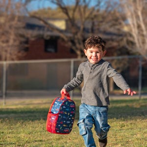 May include: A child running on grass, holding a red and blue backpack. The backpack features a pattern of vehicles and trees. The child is wearing a gray long-sleeved shirt and blue jeans.