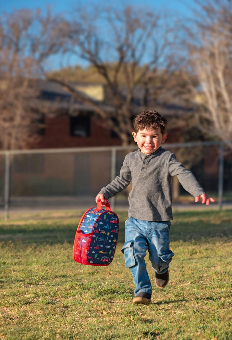 May include: A child running on grass, holding a red and blue backpack. The backpack features a pattern of vehicles and trees. The child is wearing a gray long-sleeved shirt and blue jeans.