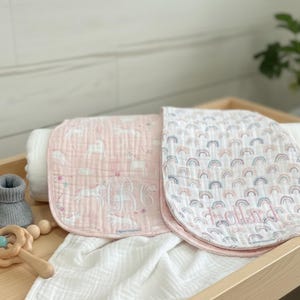 May include: A wooden tray holds baby items: two pink burp cloths with cloud and rainbow patterns, personalized with names. Gray knit baby booties and a wooden rattle with beads are also present. A white blanket is rolled up in the background.