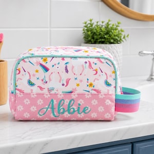 May include: A personalized cosmetic bag with a unicorn and rainbow pattern. The bag is white with pink accents and the name "Abbie" embroidered on the front. It has a teal zipper and a rainbow-striped handle.