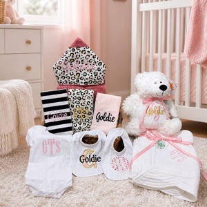 May include: A collection of baby items in a nursery setting. Includes a white teddy bear with a pink ribbon, a hooded towel with a leopard print, a white onesie, bibs, and a blanket, all personalized with names.