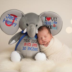 May include: A newborn baby wrapped in a cream blanket lies beside a grey plush elephant toy. The elephant is embroidered with the name "Jack Henry" and the phrase "Love you to the moon & back." The elephant also has the baby's birth stats: 3.5 kg, 8:05 pm, 51 cm.